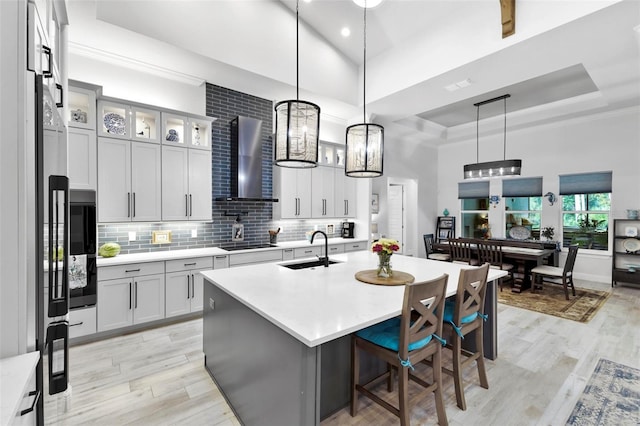 kitchen featuring sink, a tray ceiling, a center island with sink, hanging light fixtures, and wall chimney exhaust hood