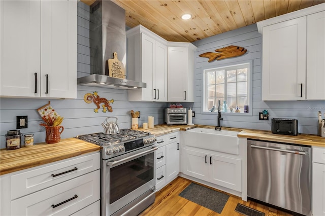 kitchen featuring stainless steel appliances, white cabinetry, sink, wooden counters, and wall chimney range hood