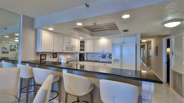 kitchen featuring a kitchen bar, kitchen peninsula, white appliances, a raised ceiling, and white cabinets