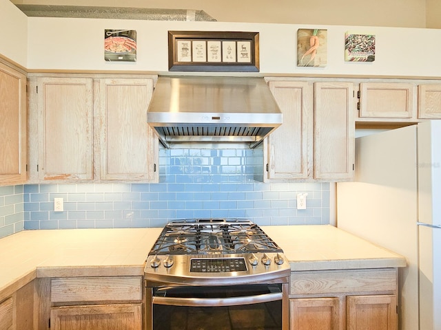 kitchen featuring backsplash, stainless steel gas range oven, wall chimney range hood, and white refrigerator