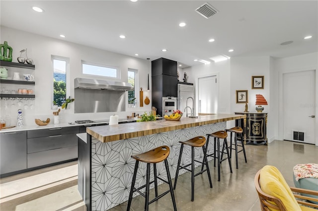 kitchen with a kitchen bar, a kitchen island with sink, wooden counters, and range hood