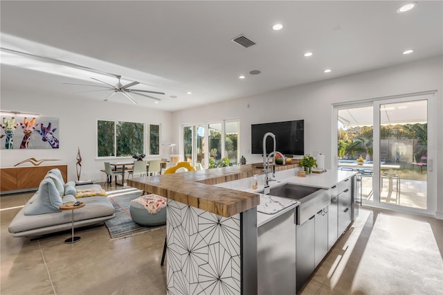 kitchen featuring wood counters, a kitchen island with sink, sink, ceiling fan, and white cabinetry