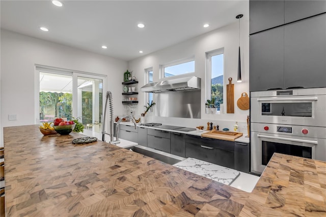 kitchen featuring wall chimney range hood, sink, double oven, decorative light fixtures, and butcher block counters