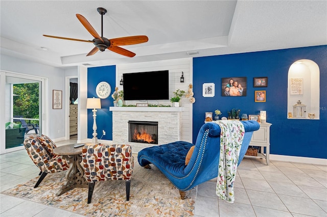 tiled living room with ceiling fan, a stone fireplace, a textured ceiling, and a tray ceiling