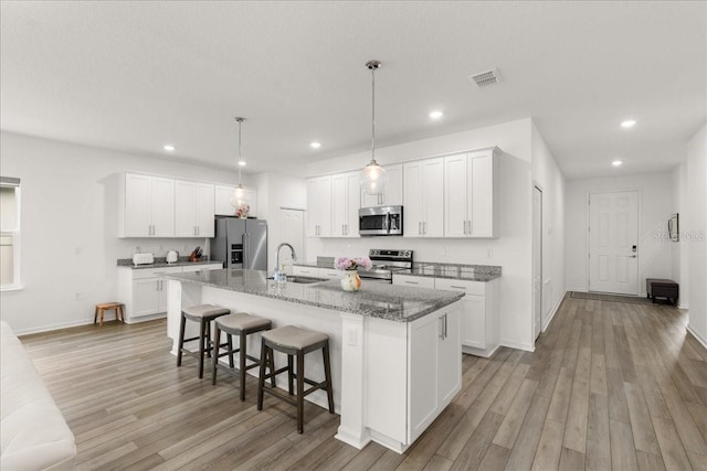 kitchen with appliances with stainless steel finishes, light hardwood / wood-style flooring, white cabinetry, hanging light fixtures, and an island with sink