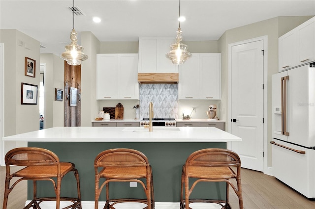 kitchen featuring backsplash, high end white refrigerator, a kitchen island with sink, white cabinets, and hanging light fixtures