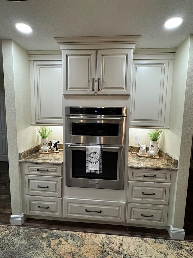 kitchen with a textured ceiling, light stone countertops, a textured wall, and stainless steel double oven