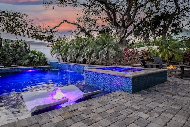 pool at dusk featuring an in ground hot tub, a fire pit, pool water feature, and a patio