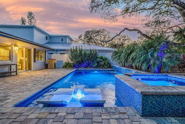 pool at dusk featuring an in ground hot tub, a patio, an outdoor kitchen, pool water feature, and an outdoor fire pit