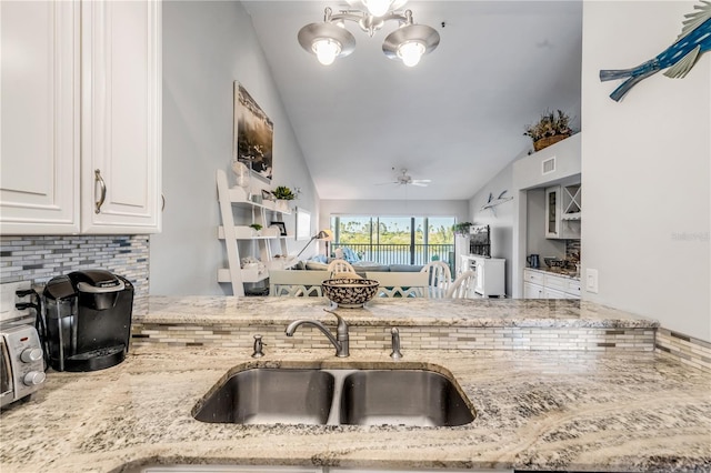 kitchen with light stone counters, ceiling fan with notable chandelier, sink, white cabinets, and lofted ceiling