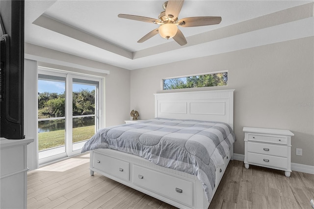 bedroom featuring ceiling fan, access to outside, a tray ceiling, a water view, and light wood-type flooring