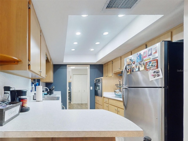 kitchen with light brown cabinets, a tray ceiling, black oven, kitchen peninsula, and stainless steel refrigerator