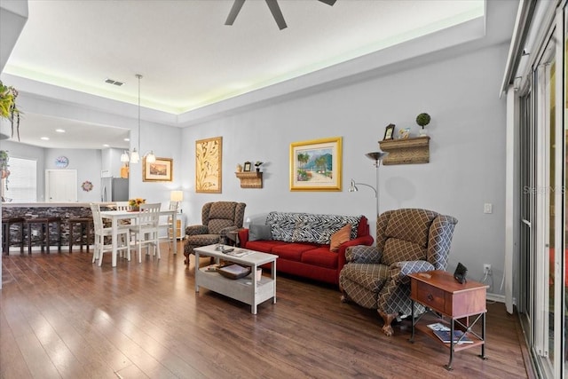 living room featuring ceiling fan with notable chandelier, a raised ceiling, and dark wood-type flooring