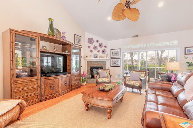 living room with ceiling fan, a tiled fireplace, vaulted ceiling, and light wood-type flooring