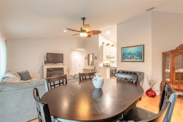 dining room with ceiling fan, a premium fireplace, light hardwood / wood-style floors, and vaulted ceiling