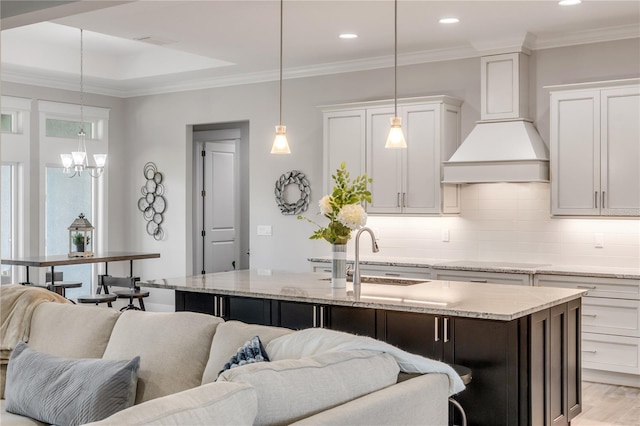 kitchen with premium range hood, white cabinetry, sink, hanging light fixtures, and a kitchen island with sink