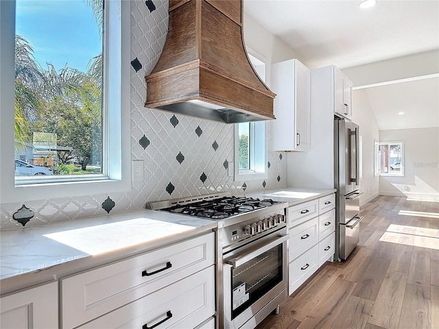 kitchen with high end appliances, white cabinetry, light stone counters, light wood-type flooring, and custom range hood