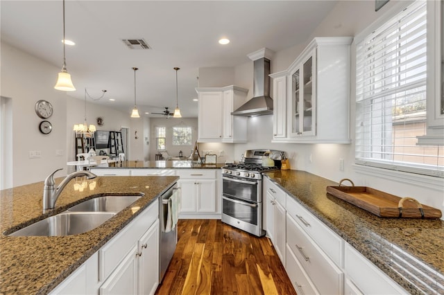 kitchen with appliances with stainless steel finishes, white cabinetry, sink, hanging light fixtures, and wall chimney range hood