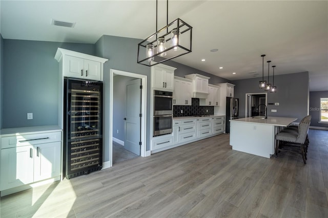 kitchen with hanging light fixtures, stainless steel appliances, wine cooler, white cabinets, and a center island with sink