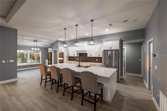 kitchen featuring appliances with stainless steel finishes, decorative backsplash, white cabinets, a center island with sink, and decorative light fixtures