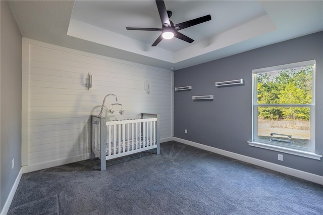 unfurnished bedroom with dark carpet, a tray ceiling, a crib, and ceiling fan