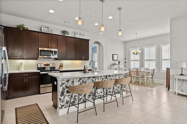 kitchen with sink, a kitchen island with sink, stainless steel appliances, tasteful backsplash, and decorative light fixtures