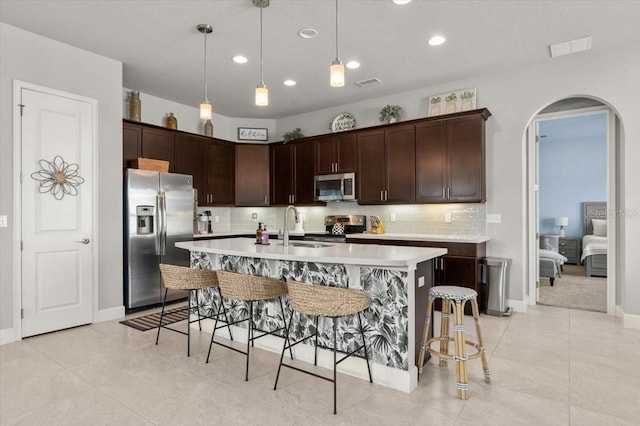 kitchen with a kitchen bar, dark brown cabinets, hanging light fixtures, appliances with stainless steel finishes, and a kitchen island with sink