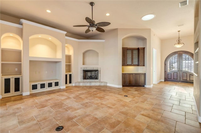 unfurnished living room featuring a tiled fireplace, built in features, french doors, and ceiling fan