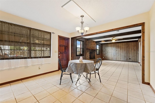 tiled dining area with a brick fireplace, a textured ceiling, wooden walls, beam ceiling, and ceiling fan with notable chandelier