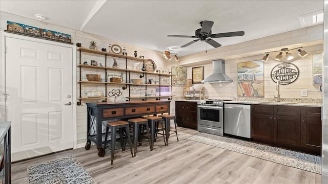 kitchen featuring appliances with stainless steel finishes, light hardwood / wood-style floors, dark brown cabinets, and wall chimney range hood