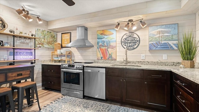 kitchen with sink, light stone counters, dark brown cabinets, appliances with stainless steel finishes, and wall chimney range hood