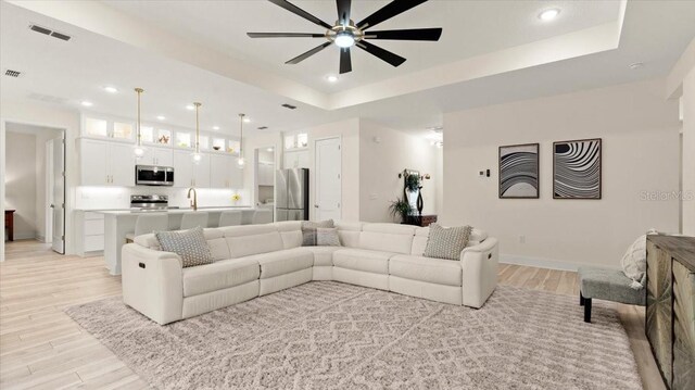 living room featuring sink, a tray ceiling, light hardwood / wood-style flooring, and ceiling fan