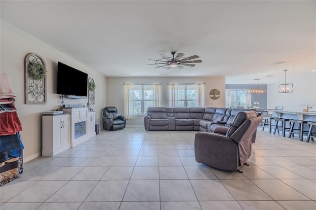 living area with baseboards, ceiling fan with notable chandelier, a glass covered fireplace, and light tile patterned flooring