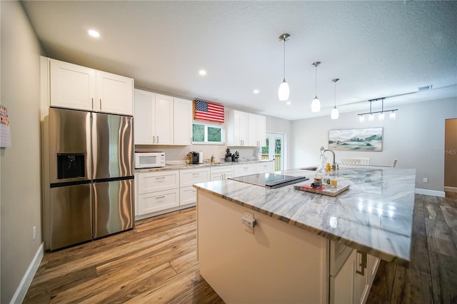 kitchen with an island with sink, black electric cooktop, pendant lighting, stainless steel fridge, and white cabinetry