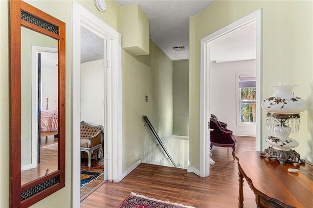 hall with a textured ceiling, wood finished floors, visible vents, an upstairs landing, and baseboards