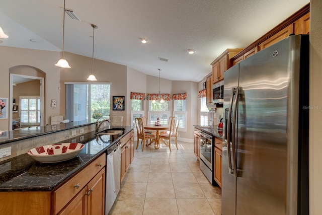 kitchen with stainless steel appliances, a sink, hanging light fixtures, brown cabinetry, and dark stone countertops