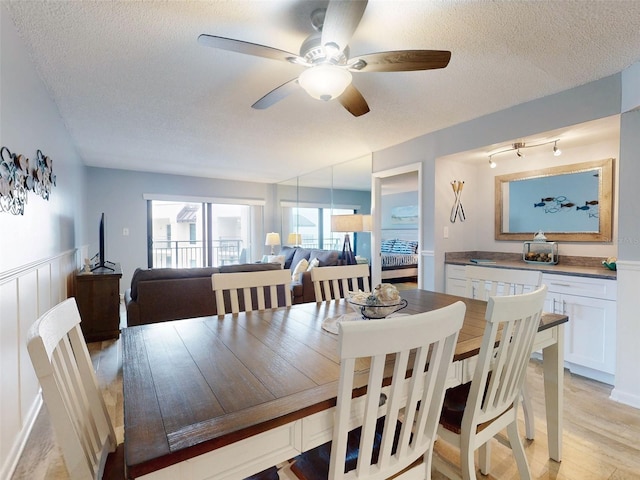 dining area with light wood finished floors, a ceiling fan, a textured ceiling, and wainscoting