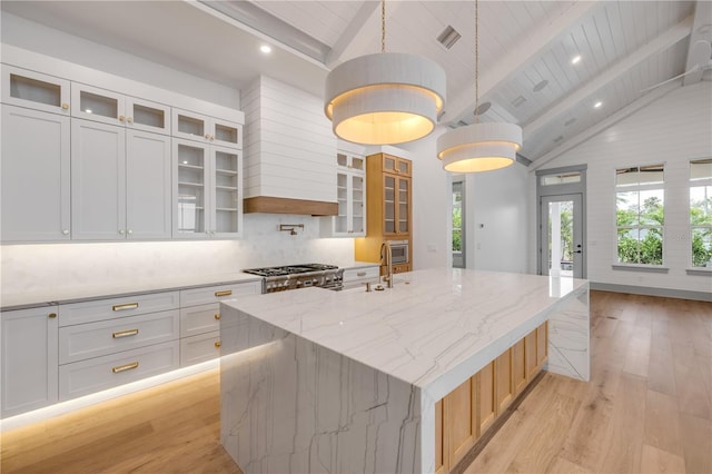 kitchen featuring stove, vaulted ceiling with beams, light stone countertops, a large island with sink, and light wood-type flooring