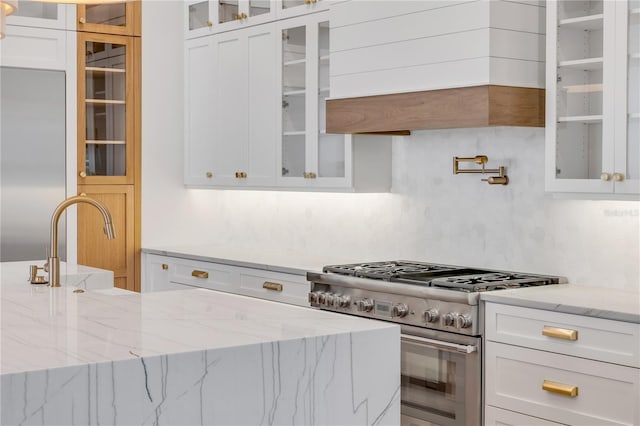 kitchen featuring light stone counters, stainless steel range, custom range hood, decorative backsplash, and white cabinets