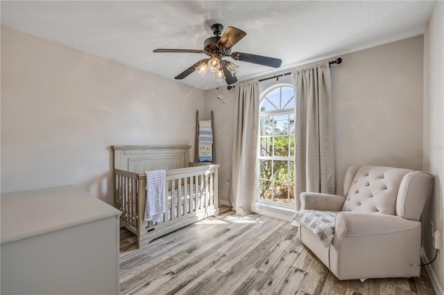 bedroom with a textured ceiling, a ceiling fan, baseboards, a nursery area, and light wood finished floors