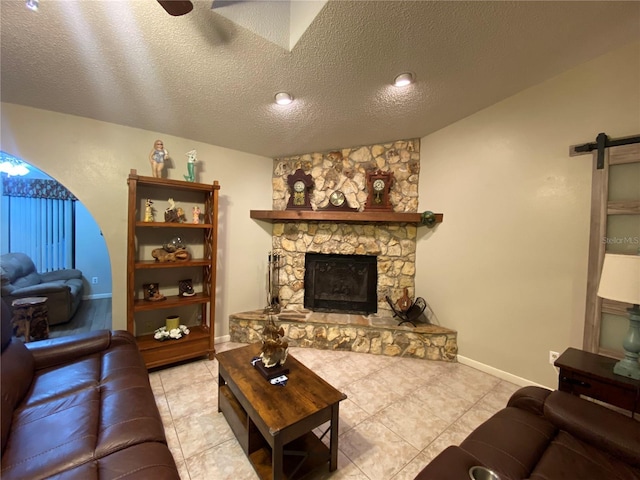 tiled living room with baseboards, a barn door, a stone fireplace, a textured ceiling, and a ceiling fan