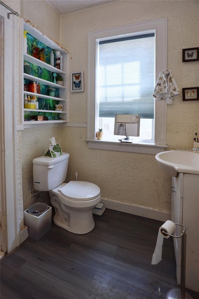 half bathroom featuring toilet, wood finished floors, baseboards, vanity, and a textured wall