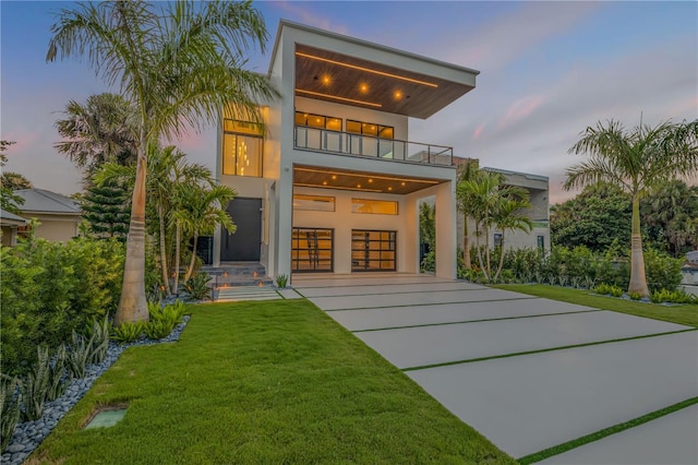 back of house with an attached garage, a balcony, a yard, concrete driveway, and stucco siding