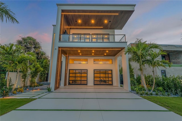 back of property at dusk with a garage, driveway, a balcony, and stucco siding