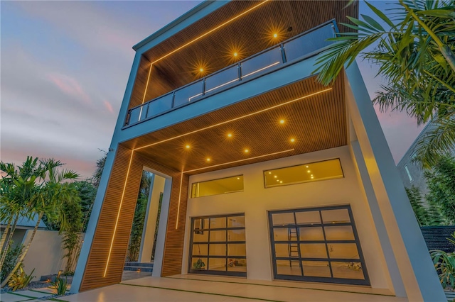 back of house at dusk featuring a balcony and stucco siding