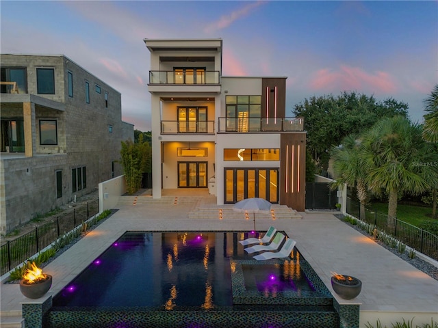 back of house at dusk with a patio, a fenced backyard, a balcony, french doors, and stucco siding