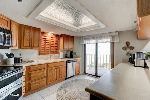 kitchen featuring brown cabinets, a sink, a tray ceiling, appliances with stainless steel finishes, and light tile patterned flooring