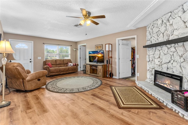 living room with a textured ceiling, visible vents, ceiling fan, wood finished floors, and a stone fireplace