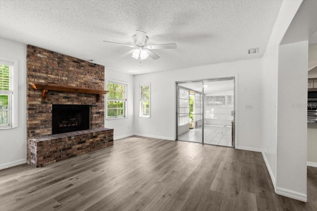unfurnished living room featuring a textured ceiling, wood finished floors, visible vents, a brick fireplace, and ceiling fan