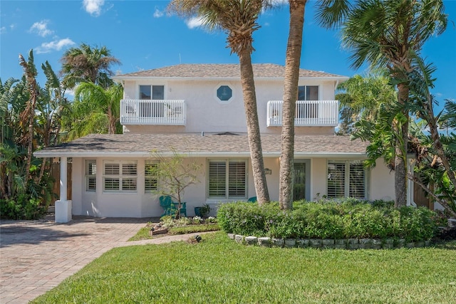 traditional-style house featuring a shingled roof, a balcony, a front yard, and stucco siding